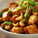Close-up of authentic cashew chicken stir-fry with golden-brown chicken cubes, toasted cashews, green onion garnish in a shallow white ceramic bowl with glossy dark amber soy-based sauce