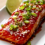 Close-up of a tender baked salmon fillet with glossy sweet chili garlic sauce, garnished with chopped green onions, sesame seeds, and a lime wedge on a white rectangular plate