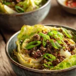 Close-up of beef cabbage bowls featuring sautéed ground beef and tender crisp cabbage leaves garnished with sliced green onions, sesame seeds, and red pepper flakes on a rustic wooden surface