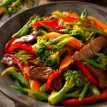 Plate of easy beef stir fry with thinly sliced beef sirloin, red and green bell peppers, carrots, broccoli florets, and green onions in a glossy sauce on a rustic wooden surface.
