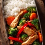 Close-up of easy chicken and vegetable stir-fry with tender chicken breast, broccoli, carrots, red bell peppers in soy garlic sauce, served with white jasmine rice in a rectangular container on rustic wooden surface
