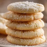 Stack of soft and chewy sugar cookies with golden edges and pale centers on a white ceramic plate, dusted with powdered sugar on a rustic wooden surface