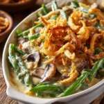Close-up of a rustic green bean casserole in a shallow oval casserole dish, showing creamy mushroom sauce, tender green beans, and crispy golden fried onions under warm natural light.