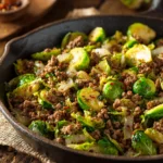 Hearty skillet of cooked ground beef and halved Brussels sprouts with diced onions and garlic, seasoned and served warm in a rustic pan on wooden surface