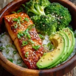 Honey glazed pan-seared salmon fillet served on fluffy jasmine rice with steamed broccoli florets, sliced avocado, sesame seeds, and chopped green onions in a rustic bowl