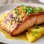 Pan-seared and oven-baked salmon fillet with a crispy golden brown exterior, brushed with bright yellow pineapple glaze, garnished with thin pineapple wedges and chopped green onions on a white plate.