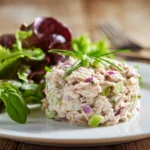Close-up of creamy classic tuna salad with chunks of tuna, finely chopped celery and red onion, served on a white round plate with bright green mixed greens on rustic wooden surface