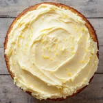 Lemon cake lemon frosting - the image is a close-up of a round cake with a golden brown crust. the cake is topped with a generous layer of white frosting that has small yellow sprinkles scattered across it. the frosting appears to be smooth and creamy, and the sprinkles add a pop of color to the cake. the background is a wooden surface, which contrasts with the warm tones of the cake and the wooden surface.