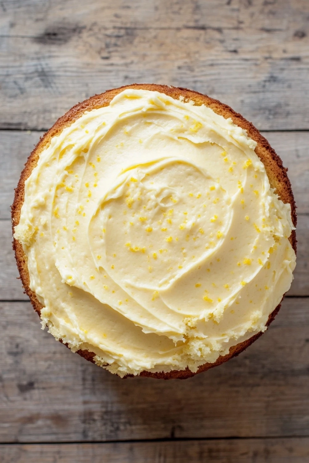 Lemon cake lemon frosting - the image is a close-up of a round cake with a golden brown crust. the cake is topped with a generous layer of white frosting that has small yellow sprinkles scattered across it. the frosting appears to be smooth and creamy, and the sprinkles add a pop of color to the cake. the background is a wooden surface, which contrasts with the warm tones of the cake and the wooden surface.